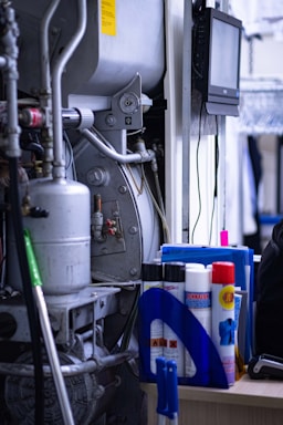 A cluttered industrial or mechanical workspace with various machines and equipment. There is a complex apparatus with pipes and metallic components, alongside a desk holding cleaning supplies, sprays, and folders. A digital screen is mounted on the wall, possibly for monitoring or control purposes.