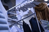 An assortment of clothing items hanging on a metal rack, including a variety of shirts in black and white. There are several wooden and metal hangers, as well as a fur coat visible in the background. The setting appears industrial, with metal pipes and beams in the upper part of the image.