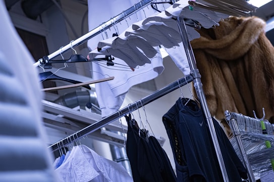 An assortment of clothing items hanging on a metal rack, including a variety of shirts in black and white. There are several wooden and metal hangers, as well as a fur coat visible in the background. The setting appears industrial, with metal pipes and beams in the upper part of the image.