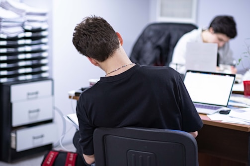 Two individuals are seated at a desk in an office setting, both working on laptops. The person in the foreground has their back to the camera, wearing a black t-shirt. The workspace appears busy, with papers, files, and other office supplies scattered across the desk. A stack of file trays is visible on the left side.