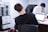 Two individuals are seated at a desk in an office setting, both working on laptops. The person in the foreground has their back to the camera, wearing a black t-shirt. The workspace appears busy, with papers, files, and other office supplies scattered across the desk. A stack of file trays is visible on the left side.