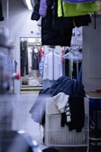A cluttered storage area with various pieces of clothing. Coats and jackets are hung on racks and hangers, with additional garments piled on a cart. The space appears narrow, with fluorescent lighting overhead.