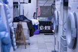 A cluttered laundry room with various garments and towels piled on a large table or basket. Several jackets and clothing items hang above, and washing machines are visible on the right. There is a chair with a fur covering, and cleaning supplies are organized on the far side near a sink.