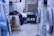 A cluttered laundry room with various garments and towels piled on a large table or basket. Several jackets and clothing items hang above, and washing machines are visible on the right. There is a chair with a fur covering, and cleaning supplies are organized on the far side near a sink.