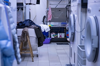 A cluttered laundry room with various garments and towels piled on a large table or basket. Several jackets and clothing items hang above, and washing machines are visible on the right. There is a chair with a fur covering, and cleaning supplies are organized on the far side near a sink.