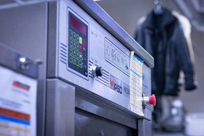 A close-up view of a commercial washing machine with a control panel, including buttons and a display. It has a printed list or instructions attached to the front. In the background, there's a blurred hanging black jacket.