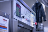 A close-up view of a commercial washing machine with a control panel, including buttons and a display. It has a printed list or instructions attached to the front. In the background, there's a blurred hanging black jacket.