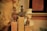 Three ancient-style figurines are displayed on wooden pedestals. The figures have intricate details and varying shapes. In the background, a white bust of a uniformed person with a hat is visible. A reddish-brown clay tablet hangs on the wall, inscribed with what seems like ancient writing.