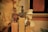 Three ancient-style figurines are displayed on wooden pedestals. The figures have intricate details and varying shapes. In the background, a white bust of a uniformed person with a hat is visible. A reddish-brown clay tablet hangs on the wall, inscribed with what seems like ancient writing.