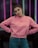 A person stands confidently with hands on hips, wearing a pink sweatshirt with a motivational question printed on it and blue jeans. The background features vertical neon lights in various colors, creating a vibrant and modern setting.
