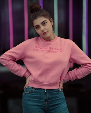 A person stands confidently with hands on hips, wearing a pink sweatshirt with a motivational question printed on it and blue jeans. The background features vertical neon lights in various colors, creating a vibrant and modern setting.
