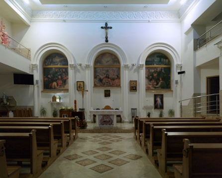A well-lit church interior with rows of wooden pews leading up to an ornate altar. The altar is adorned with religious paintings depicting biblical scenes and a crucifix hangs above. The walls feature arches and columns, and there are decorative elements like flowers and candlesticks.