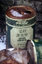 A rusted can of rapeseed cooking oil is partially covered with snow, indicating it has been outdoors for some time. The can appears aged, with text in multiple languages and an overall worn appearance. In the foreground, there is another partially visible, similarly rusted can.