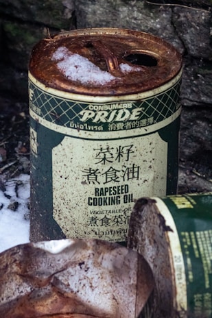 A rusted can of rapeseed cooking oil is partially covered with snow, indicating it has been outdoors for some time. The can appears aged, with text in multiple languages and an overall worn appearance. In the foreground, there is another partially visible, similarly rusted can.