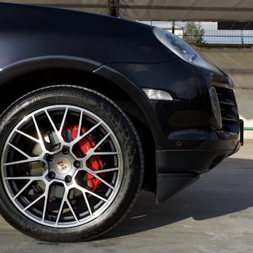 A close-up view of the front wheel of a luxury car, showcasing the intricate design of the alloy wheel with a visible logo at the center. The brake calipers painted in red are prominently visible behind the wheel spokes. The car's body is a sleek dark color, reflecting light subtly. The background is an out-of-focus area with a metal fence and part of a concrete structure, suggesting an outdoor setting.