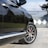 A close-up view of a luxury car parked on a cement driveway, highlighting the detailed design of its alloy wheel with red brake calipers. The car has a sleek black finish reflecting nearby greenery and shadows. In the background, a geometric patterned gate and some trees are visible.