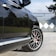 A close-up view of a luxury car parked on a cement driveway, highlighting the detailed design of its alloy wheel with red brake calipers. The car has a sleek black finish reflecting nearby greenery and shadows. In the background, a geometric patterned gate and some trees are visible.