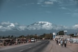 A scenic view of a snow-capped mountain in the background with a road leading towards it. On one side of the road, there are small market stalls with vendors and pedestrians. A few people are walking and carrying items, while vehicles pass by on the road. The sky is partly cloudy, adding to the serene environment.