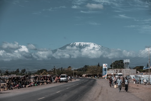 A scenic view of a snow-capped mountain in the background with a road leading towards it. On one side of the road, there are small market stalls with vendors and pedestrians. A few people are walking and carrying items, while vehicles pass by on the road. The sky is partly cloudy, adding to the serene environment.