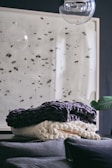 A cozy, textured blanket is artfully arranged on a dark surface, with a modern wall art piece featuring black specks in the background. A large, transparent glass orb hangs above, adding a reflective quality and contemporary feel to the space.