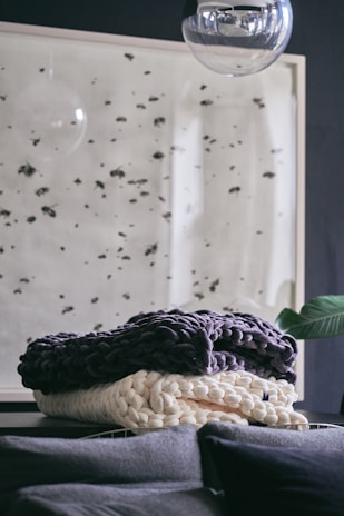 A cozy, textured blanket is artfully arranged on a dark surface, with a modern wall art piece featuring black specks in the background. A large, transparent glass orb hangs above, adding a reflective quality and contemporary feel to the space.