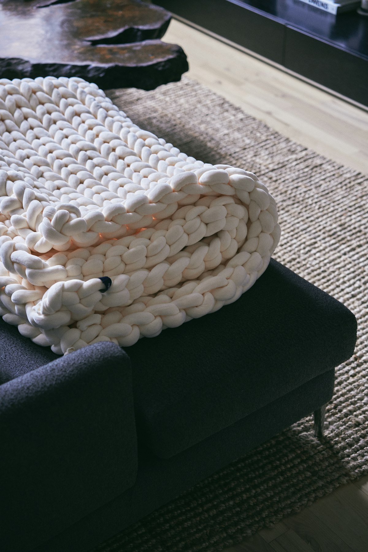 A heavy cream knitted blanket folded over the arm of a dark grey sofa