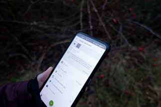 A person is holding a smartphone displaying an app or webpage against a blurred background of outdoor foliage with visible small red berries. The phone screen has text and icons related to an observation or note-taking application.