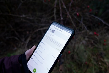 A person is holding a smartphone displaying an app or webpage against a blurred background of outdoor foliage with visible small red berries. The phone screen has text and icons related to an observation or note-taking application.