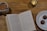 A cozy setting on a wooden table features an open book with visible text, a mug filled with tea or coffee with a spoon inside, a lit candle in a glass holder, and a plate containing two chocolate truffles. The dim lighting creates a warm, inviting atmosphere.