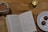 A cozy setting on a wooden table features an open book with visible text, a mug filled with tea or coffee with a spoon inside, a lit candle in a glass holder, and a plate containing two chocolate truffles. The dim lighting creates a warm, inviting atmosphere.