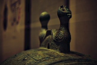A close-up view of an ornate, ancient-looking bronze artifact featuring intricate carvings and two stylized bird-like figures on top. The image highlights the details and patina of the bronze, suggesting age and historical significance.