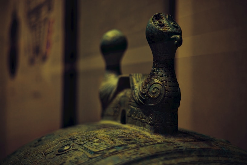 A close-up view of an ornate, ancient-looking bronze artifact featuring intricate carvings and two stylized bird-like figures on top. The image highlights the details and patina of the bronze, suggesting age and historical significance.