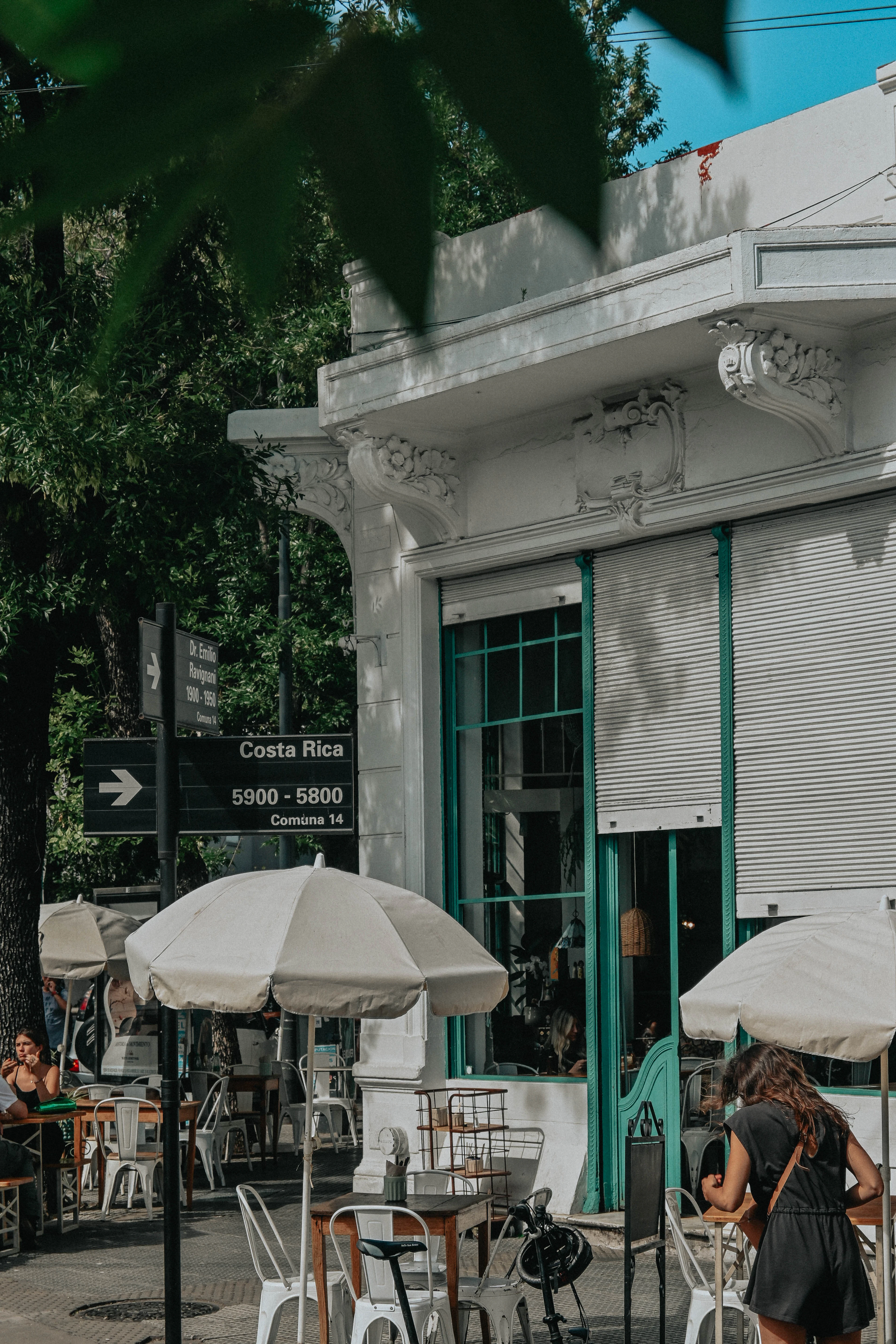 An ornate white corner building with outdoor umbrellas and diners on a tree-lined street.