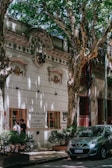 A tree-lined street features a historic building with ornate architectural details and a sign for 'Sonia Williams, Disfraces Vestuario'. Two people are standing by a window of the building, and a small car is parked on the street surrounded by lush, green foliage.