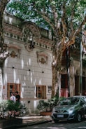 A tree-lined street features a historic building with ornate architectural details and a sign for 'Sonia Williams, Disfraces Vestuario'. Two people are standing by a window of the building, and a small car is parked on the street surrounded by lush, green foliage.