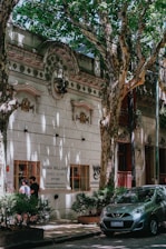 A tree-lined street features a historic building with ornate architectural details and a sign for 'Sonia Williams, Disfraces Vestuario'. Two people are standing by a window of the building, and a small car is parked on the street surrounded by lush, green foliage.