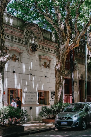 A tree-lined street features a historic building with ornate architectural details and a sign for 'Sonia Williams, Disfraces Vestuario'. Two people are standing by a window of the building, and a small car is parked on the street surrounded by lush, green foliage.