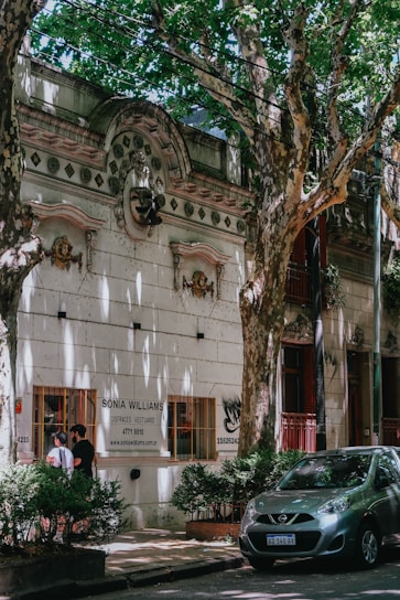 A tree-lined street features a historic building with ornate architectural details and a sign for 'Sonia Williams, Disfraces Vestuario'. Two people are standing by a window of the building, and a small car is parked on the street surrounded by lush, green foliage.