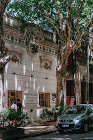 A tree-lined street features a historic building with ornate architectural details and a sign for 'Sonia Williams, Disfraces Vestuario'. Two people are standing by a window of the building, and a small car is parked on the street surrounded by lush, green foliage.