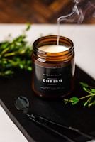 A brown glass candle jar labeled 'CHRISM' contains a white candle with wisps of smoke rising elegantly. Green sprigs and a black candle accessory are placed artistically around it on a dark surface, adding to the composition.