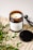 A lit scented candle in a brown glass jar with a label reading 'Verso L’Alto' is placed on a textured beige surface. Fresh green foliage is scattered around the candle, and a black metal lid and wick trimmer are positioned nearby.