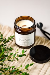 A lit scented candle in a brown glass jar with a label reading 'Verso L’Alto' is placed on a textured beige surface. Fresh green foliage is scattered around the candle, and a black metal lid and wick trimmer are positioned nearby.