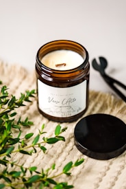 A lit scented candle in a brown glass jar with a label reading 'Verso L’Alto' is placed on a textured beige surface. Fresh green foliage is scattered around the candle, and a black metal lid and wick trimmer are positioned nearby.