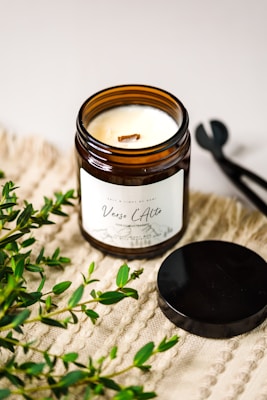 A lit scented candle in a brown glass jar with a label reading 'Verso L’Alto' is placed on a textured beige surface. Fresh green foliage is scattered around the candle, and a black metal lid and wick trimmer are positioned nearby.
