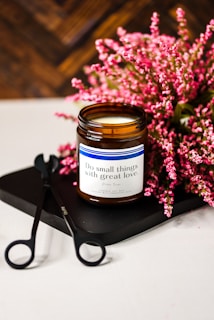 A brown glass jar candle sits on a black tray next to a pair of wick trimmers. In the background, clusters of pink flowers add a touch of color. The candle label reads 'Do small things with great love.' The setting conveys a sense of care and tranquility.