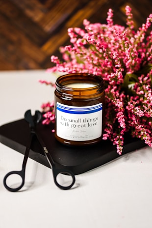 A brown glass jar candle sits on a black tray next to a pair of wick trimmers. In the background, clusters of pink flowers add a touch of color. The candle label reads 'Do small things with great love.' The setting conveys a sense of care and tranquility.