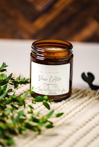 A brown glass jar candle with a white label named 'Verso L'Alto' is placed on a textured beige surface. Green leafy branches are positioned beside the jar, adding a natural element. The background is a warm, out-of-focus wooden texture.