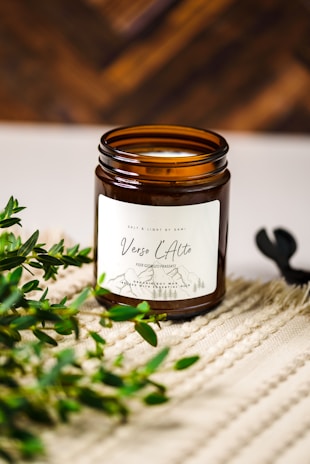 A brown glass jar candle with a white label named 'Verso L'Alto' is placed on a textured beige surface. Green leafy branches are positioned beside the jar, adding a natural element. The background is a warm, out-of-focus wooden texture.