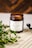 A brown glass jar candle with a white label named 'Verso L'Alto' is placed on a textured beige surface. Green leafy branches are positioned beside the jar, adding a natural element. The background is a warm, out-of-focus wooden texture.