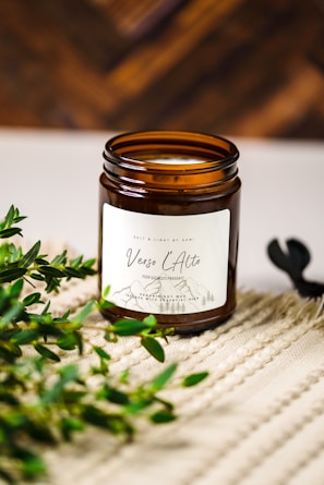 A brown glass jar candle with a white label named 'Verso L'Alto' is placed on a textured beige surface. Green leafy branches are positioned beside the jar, adding a natural element. The background is a warm, out-of-focus wooden texture.